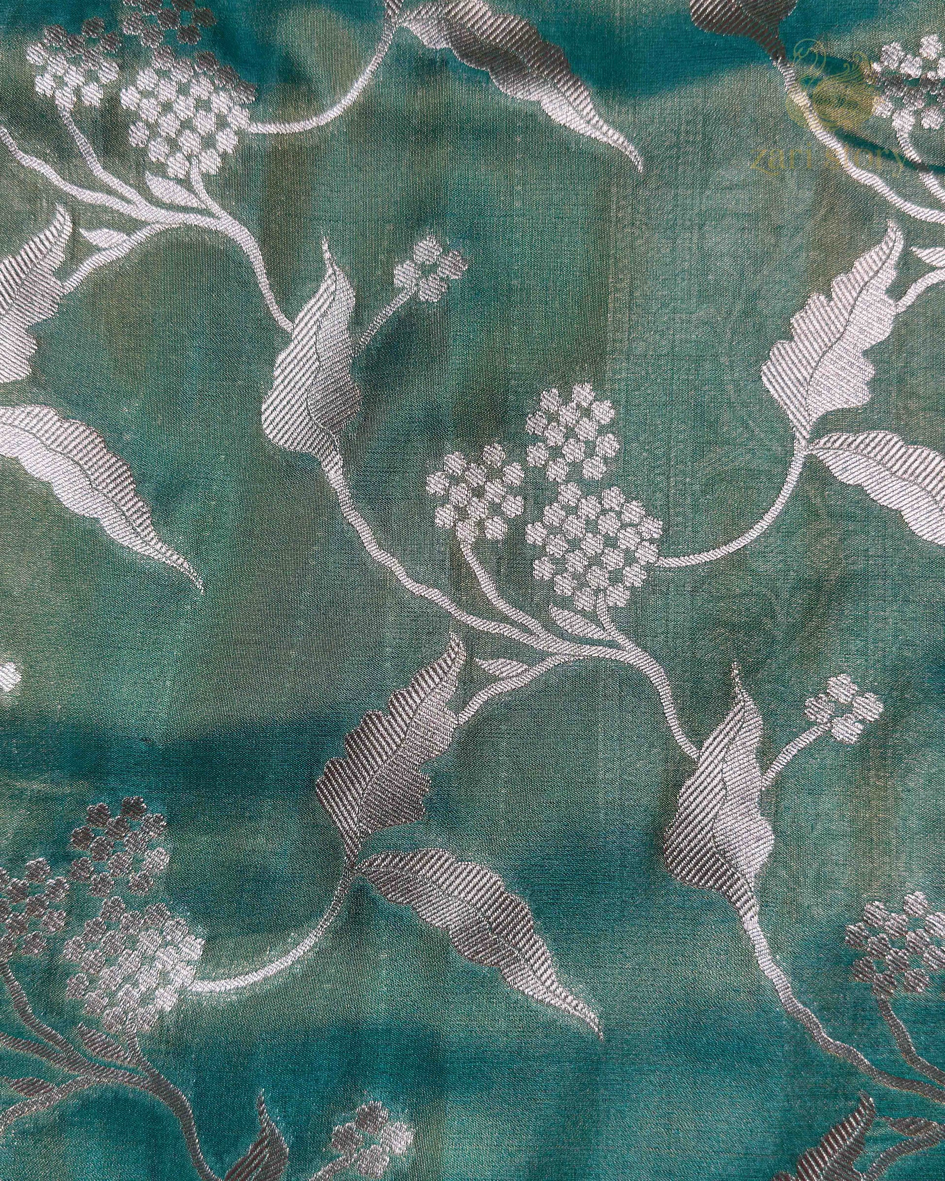 A close-up of teal green fabric with a woven floral pattern in light gray and white.