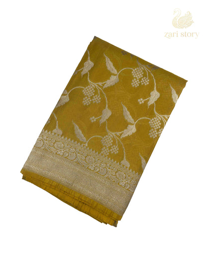 A folded yellow-gold saree with intricate silver floral and vine motifs, displayed on a clean white background with a 'zari story' logo in the top right corner.