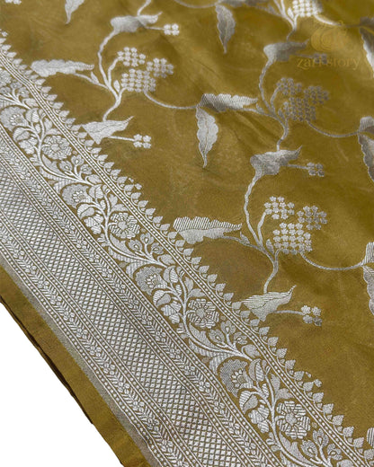 A close-up of a mustard yellow or ochre-colored fabric with intricate silver floral and bird motifs woven across it, featuring a wide decorative silver border.