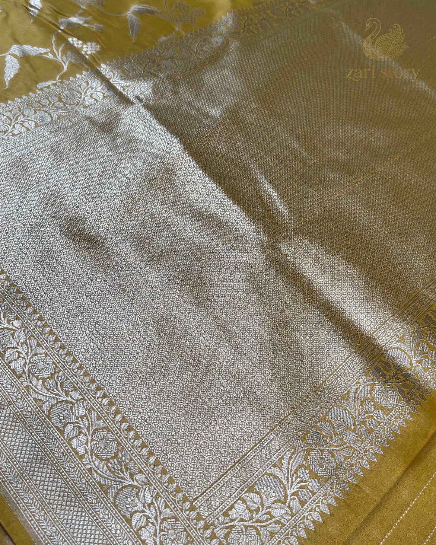 A close-up of a woven textile, likely a sari, featuring a large silver-toned central panel with intricate floral and geometric motifs, bordered by a matching teal-green border with floral embroidery and a small gold zari story logo in the top right.