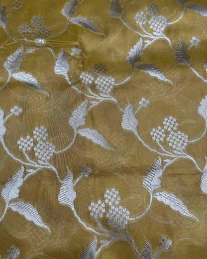 A close-up of a gold-colored sheer fabric with a silver floral and vine pattern woven across it.